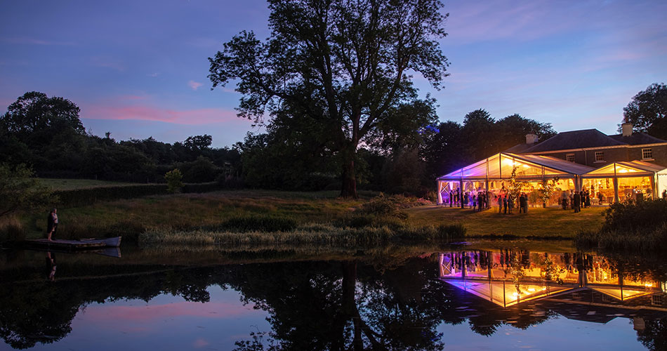 A stunning clear span marquee at night as the sun sets, next to a lake in a private house. It is busy with people and coloured lights from the marquee are reflected in the water of the lake.