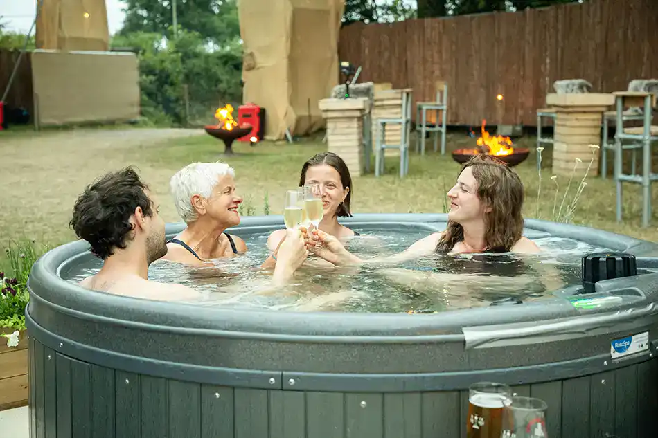 Festival guests raising a champagne toast while relaxing in the wood-clad hot tubs at the VIP camping spa area for glamping Glastonbury 2027.