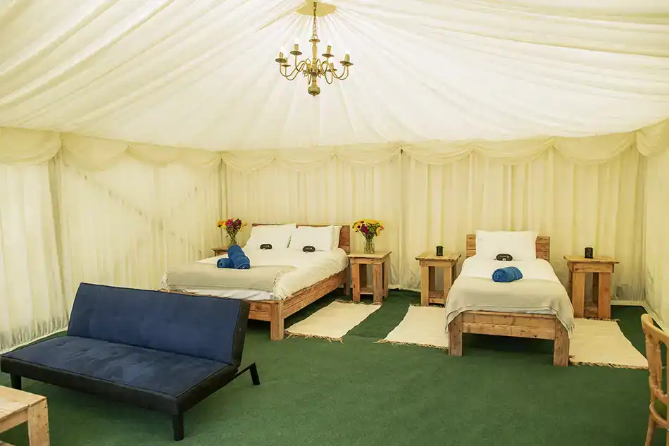The spacious, carpeted interior of a safari tent featuring a double bed, single bed, sofa, rustic side tables, and a chandelier.