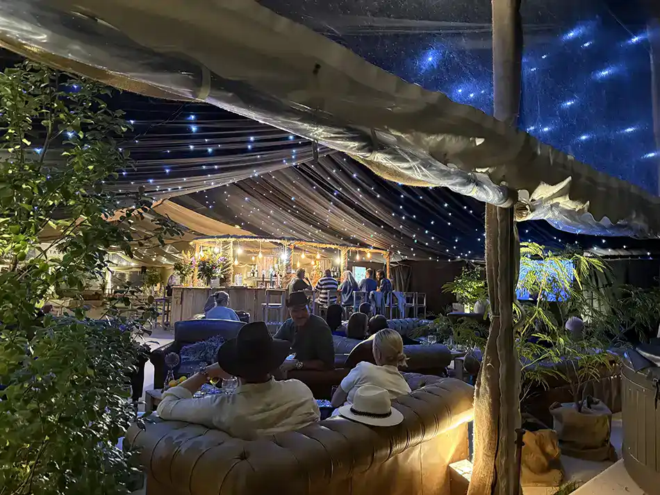 Guests relaxing on leather Chesterfield sofas under fairy lights at the vibrant lounge bar during a luxury camping at Glastonbury festival weekend.